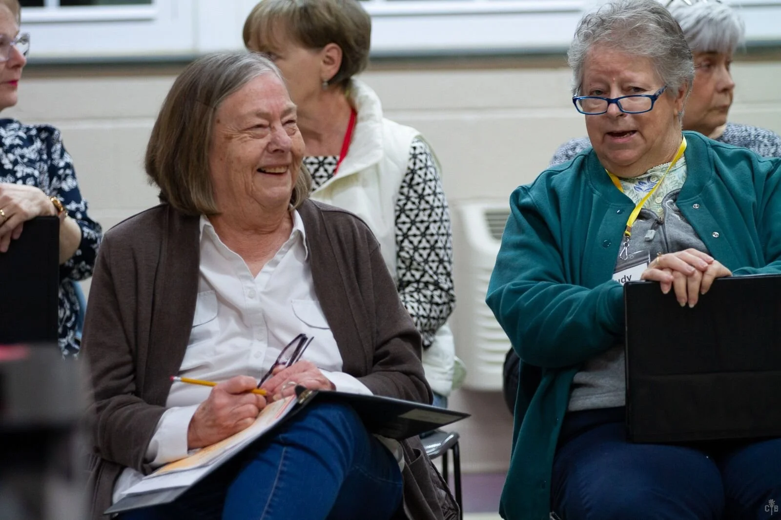 Two elderly women smiling and talking during a gathering, one taking notes with a pencil and the other holding a laptop, in an indoor setting.