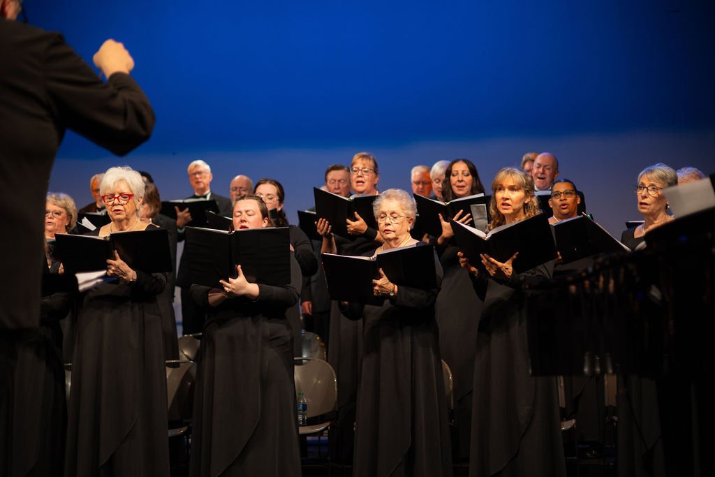 A choir of predominantly older women and men singing during a performance, with the conductor visible from behind in the foreground.
