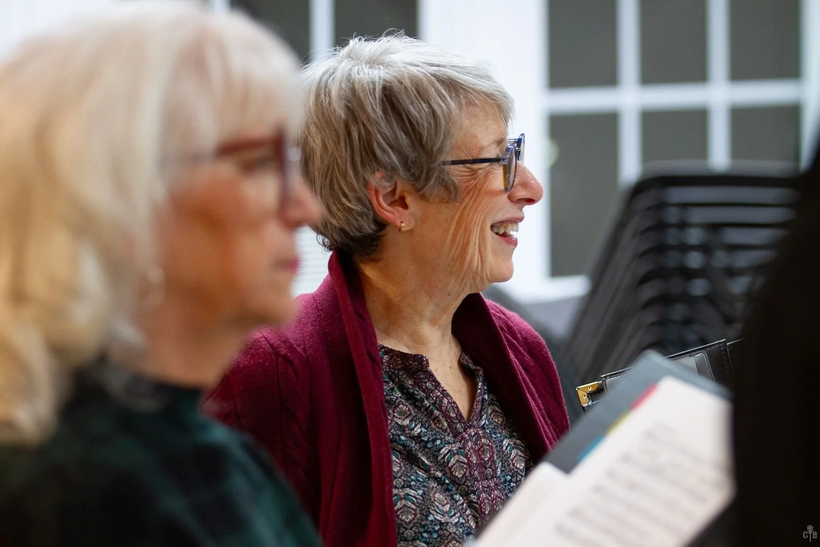 Two older women sitting in a row at a table, smiling and reading documents, with a background of stacked chairs and windows.
