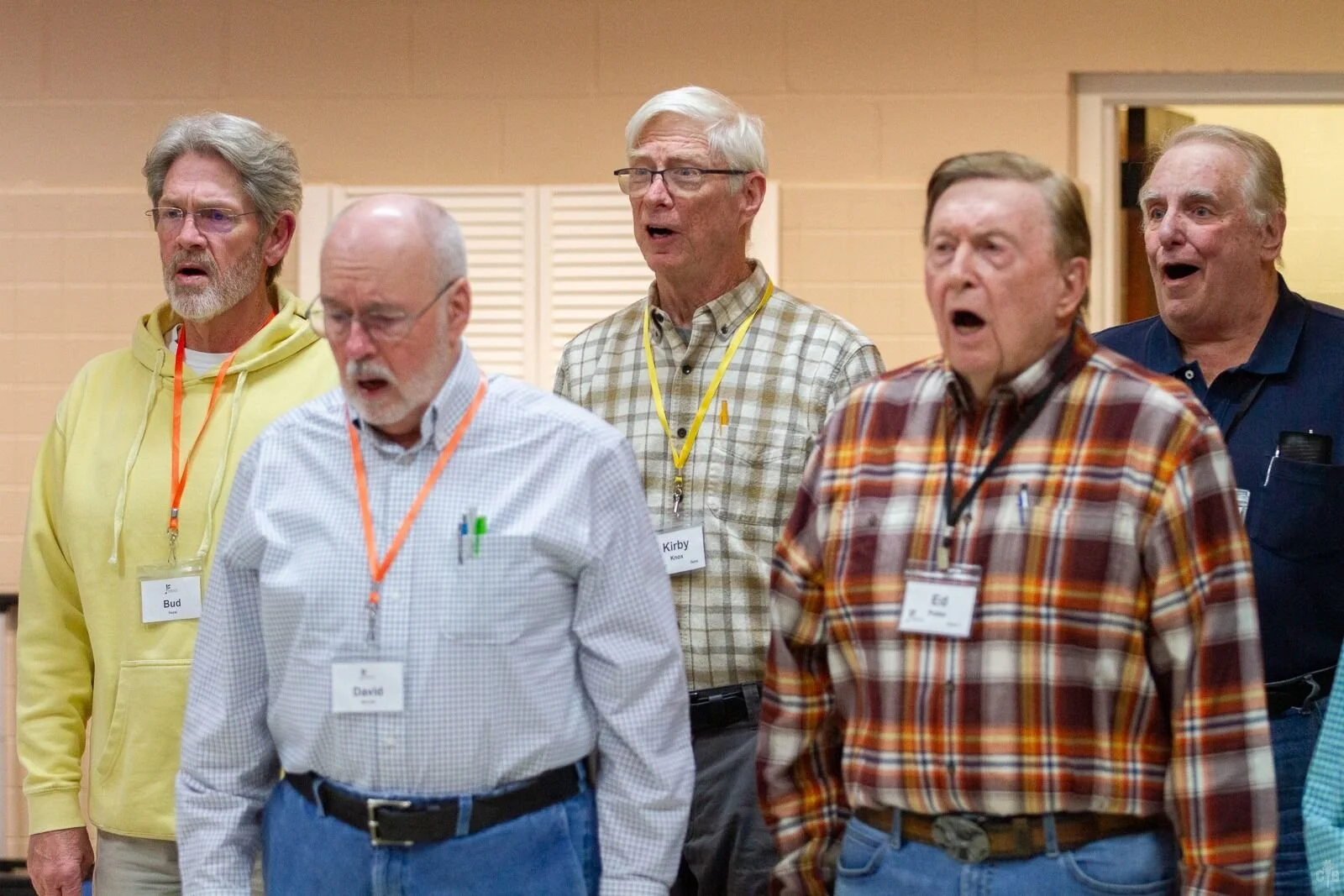 Six older men singing together at an indoor gathering, wearing casual clothing and name tags.