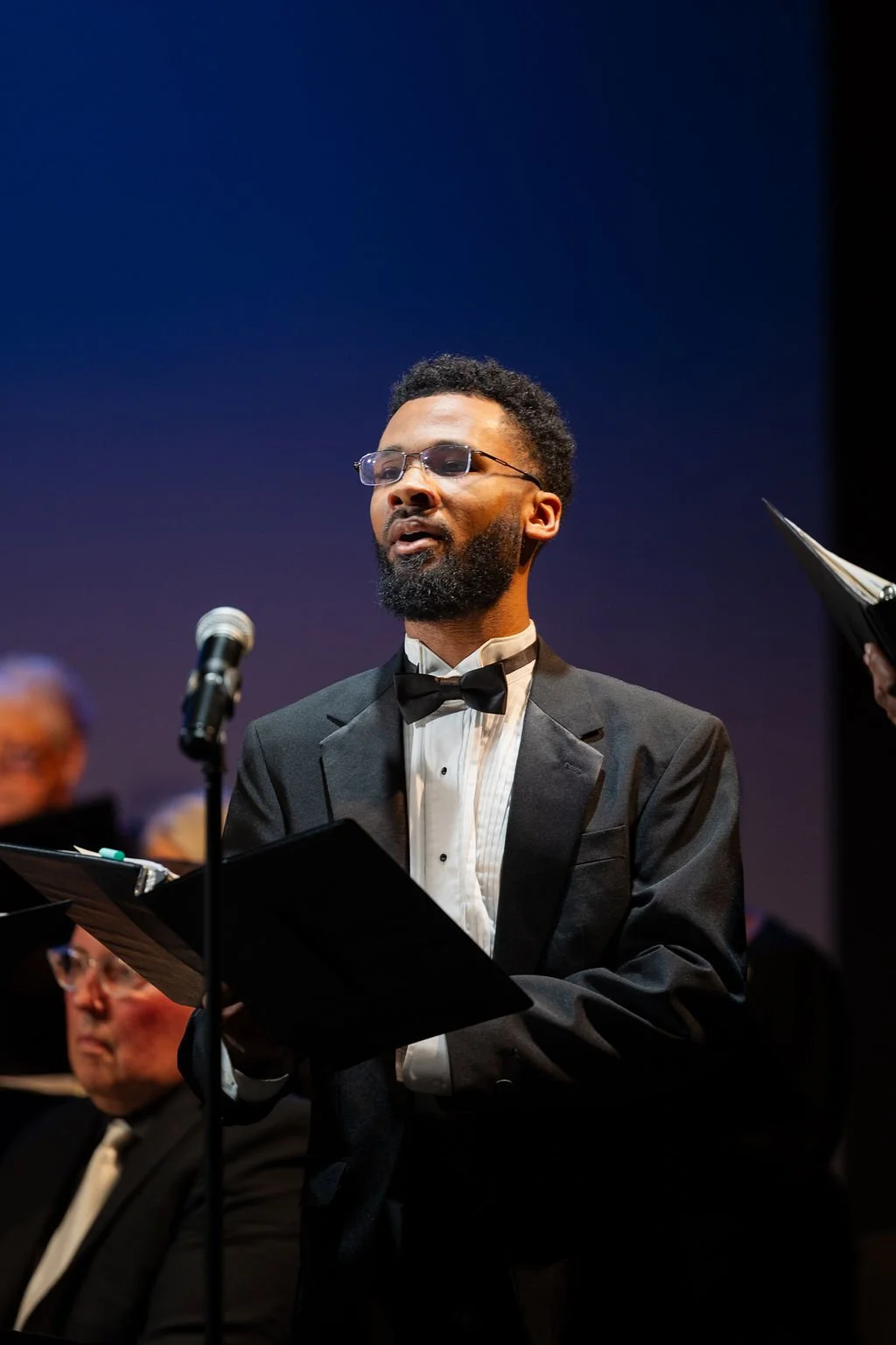 A man singing or speaking into a microphone while holding sheet music, dressed in a tuxedo with a bow tie, performing with an orchestra.