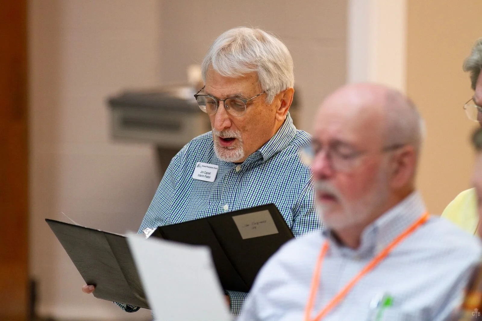 Older man with gray hair and glasses singing in a choir, holding a black folder with sheet music, wearing a green checkered shirt and name tag. Other choir members are visible in the background.