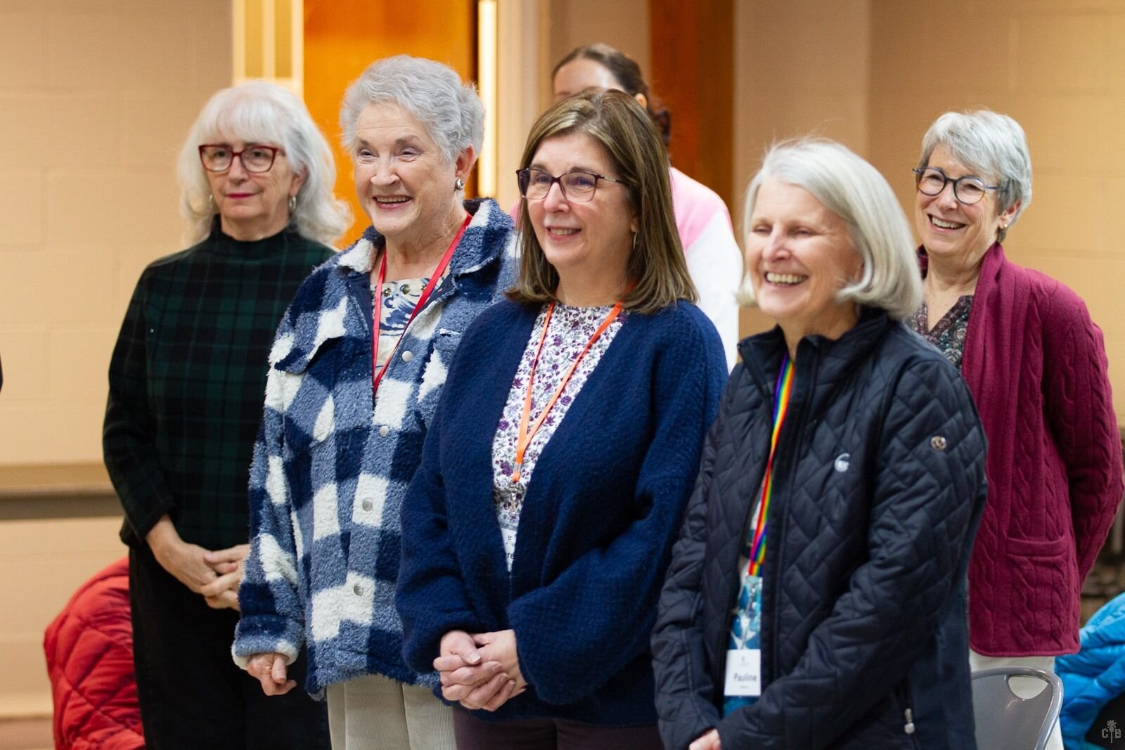 Group of six women standing indoors, smiling and looking towards the front, at a social or professional event.