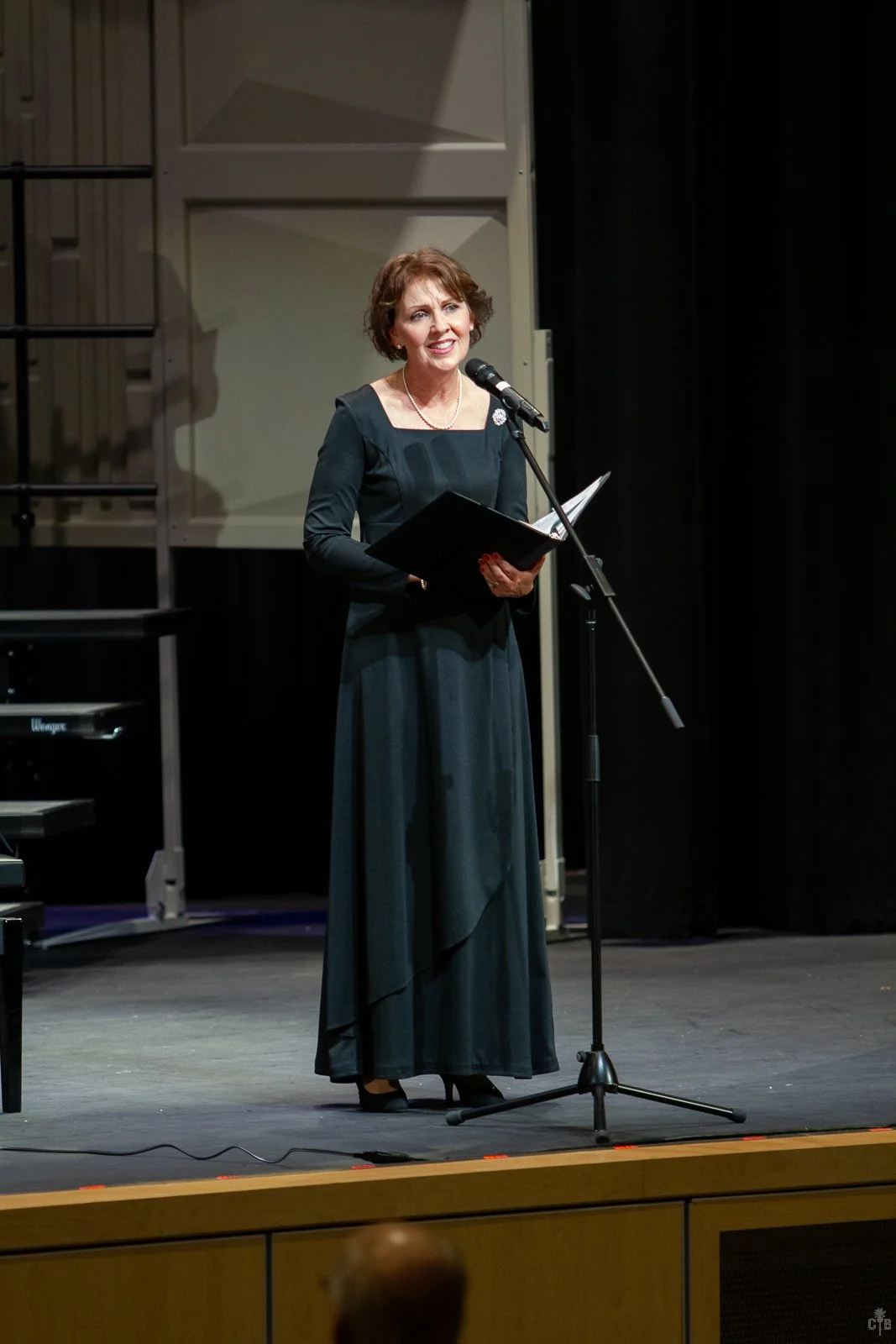 A woman in a long dark dress stands on stage, speaking into a microphone and holding a folder, with a music stand and piano behind her.