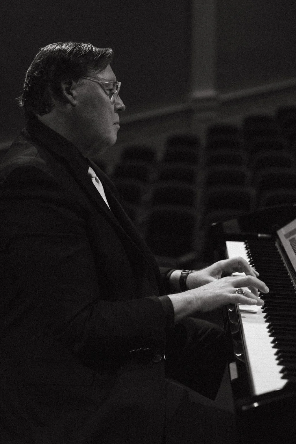 A person with short hair, glasses, and a watch is playing a piano in a dimly lit room, facing the keyboard with sheet music nearby.