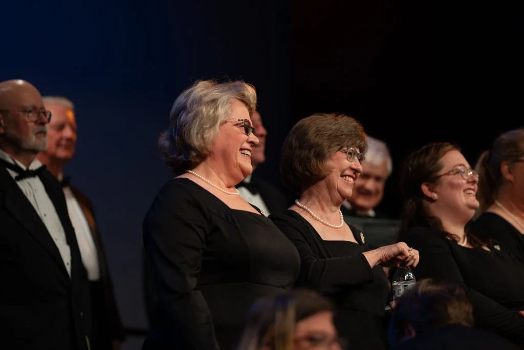A group of people dressed formally, mostly women in black dresses and pearl necklaces, smiling and laughing at an event.