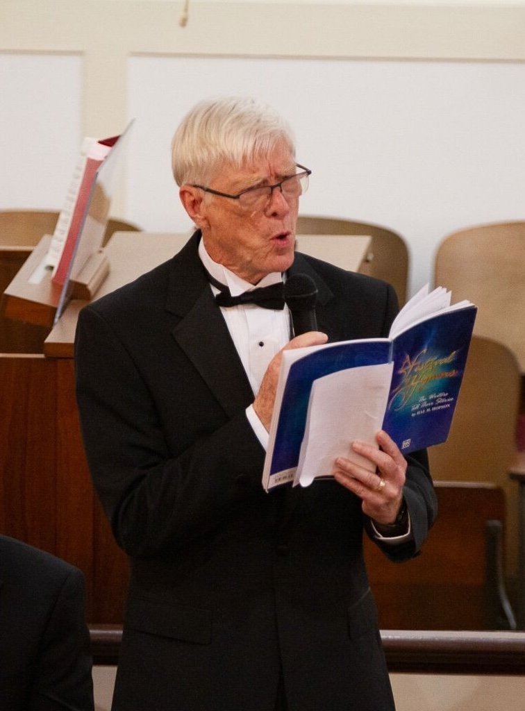 An elderly man in a tuxedo with glasses holding a microphone in one hand and a book in the other, singing or reading aloud in a church or similar setting.
