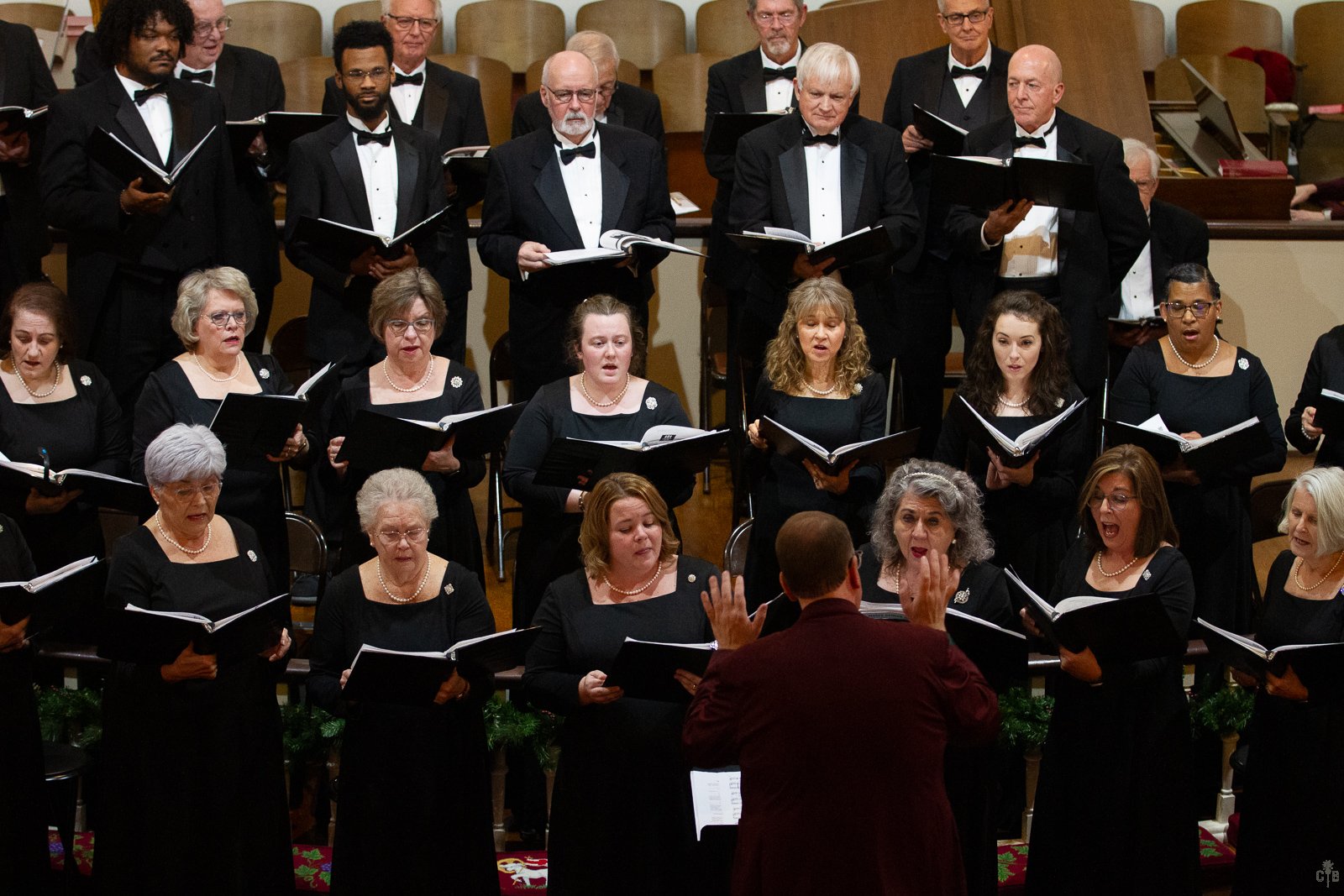 A choir performing on stage with a conductor facing them, dressed in formal black attire. The choir members are holding sheet music and singing, with a background of empty seats.