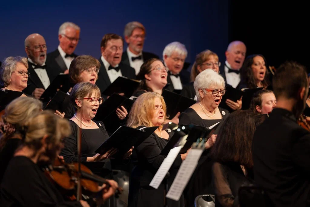 A choir of diverse older men and women singing in a concert hall, accompanied by musicians playing string instruments.
