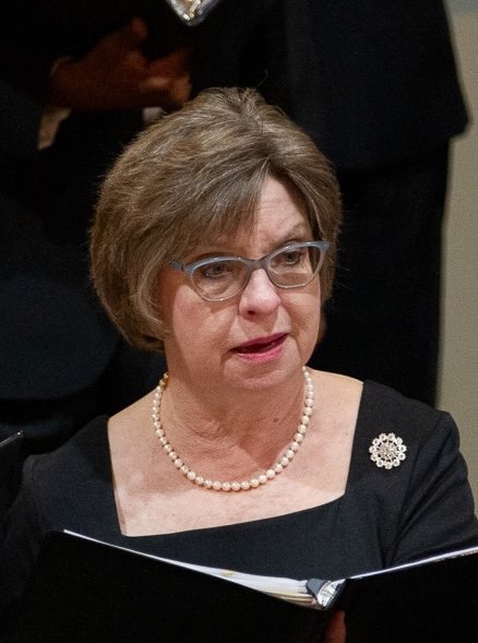 A woman with short gray hair and glasses, wearing a black dress with a pearl necklace and a decorative pin, reading a document.