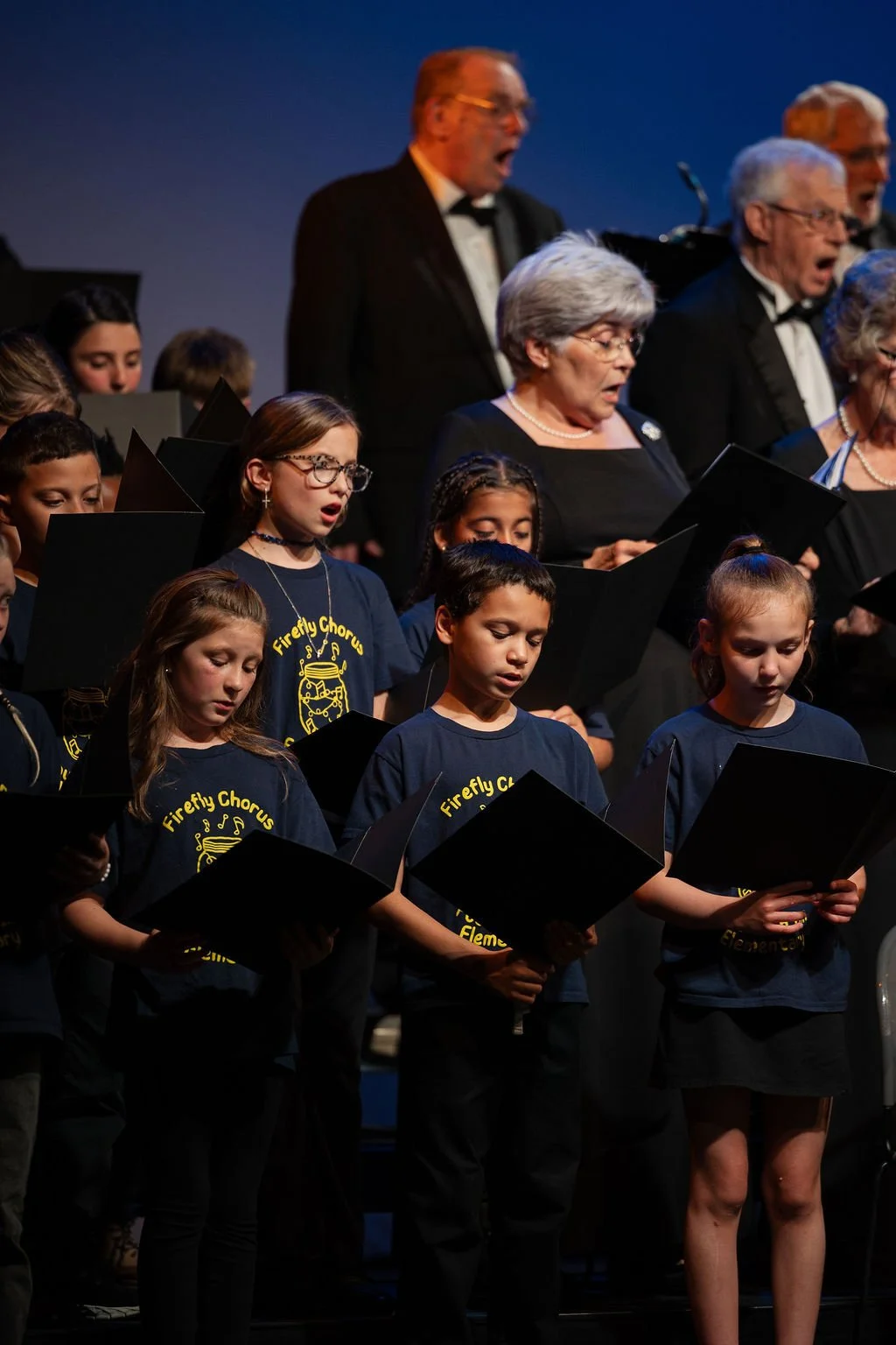 A choir of children and adults singing on stage, some wearing navy blue shirts with yellow text, in a concert setting.