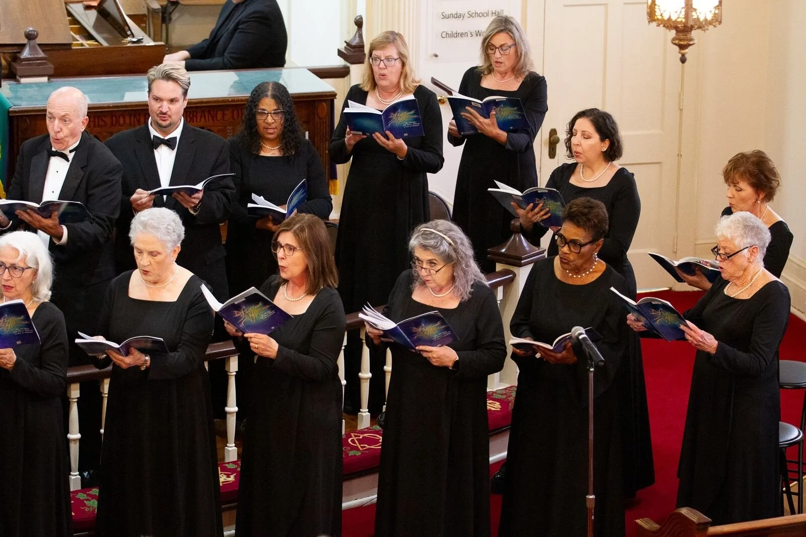 A choir of men and women dressed in black, singing from sheet music inside a church or concert hall.