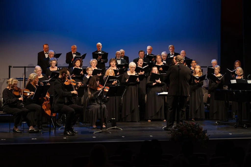 A choir and orchestra performing on stage with a conductor directing them, in front of a blue backdrop.