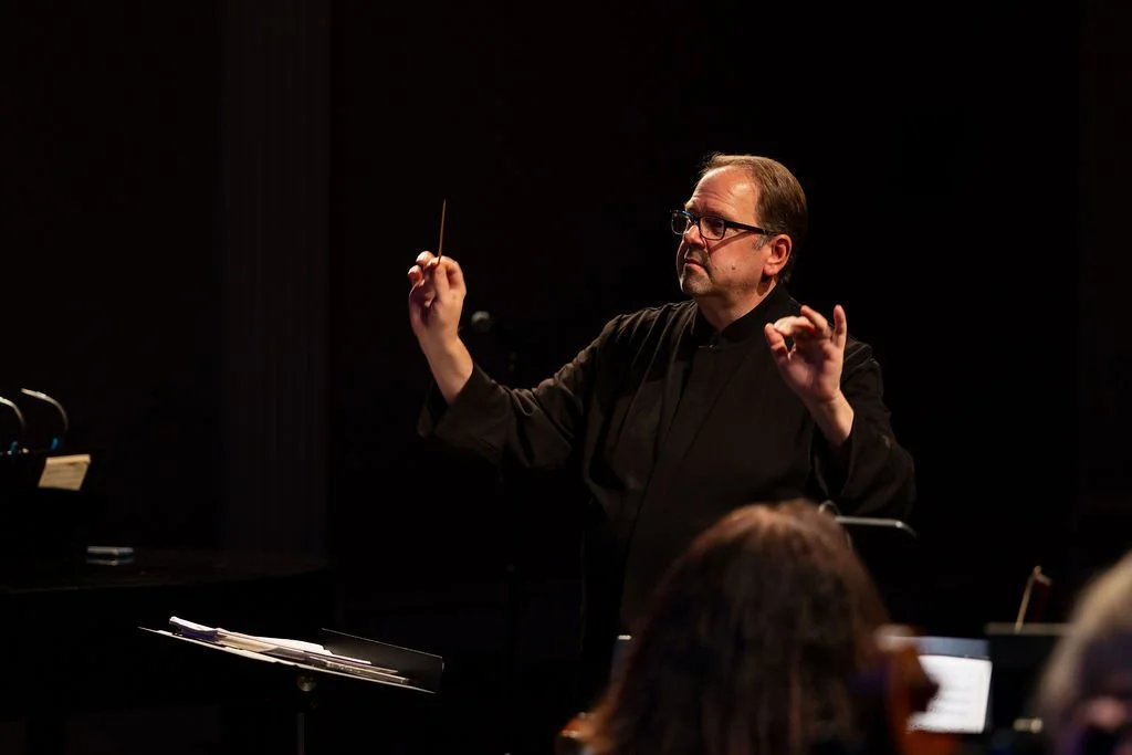 A man conducting an orchestra or choir on stage, holding a baton, with a music stand and sheet music in front of him, against a dark background.