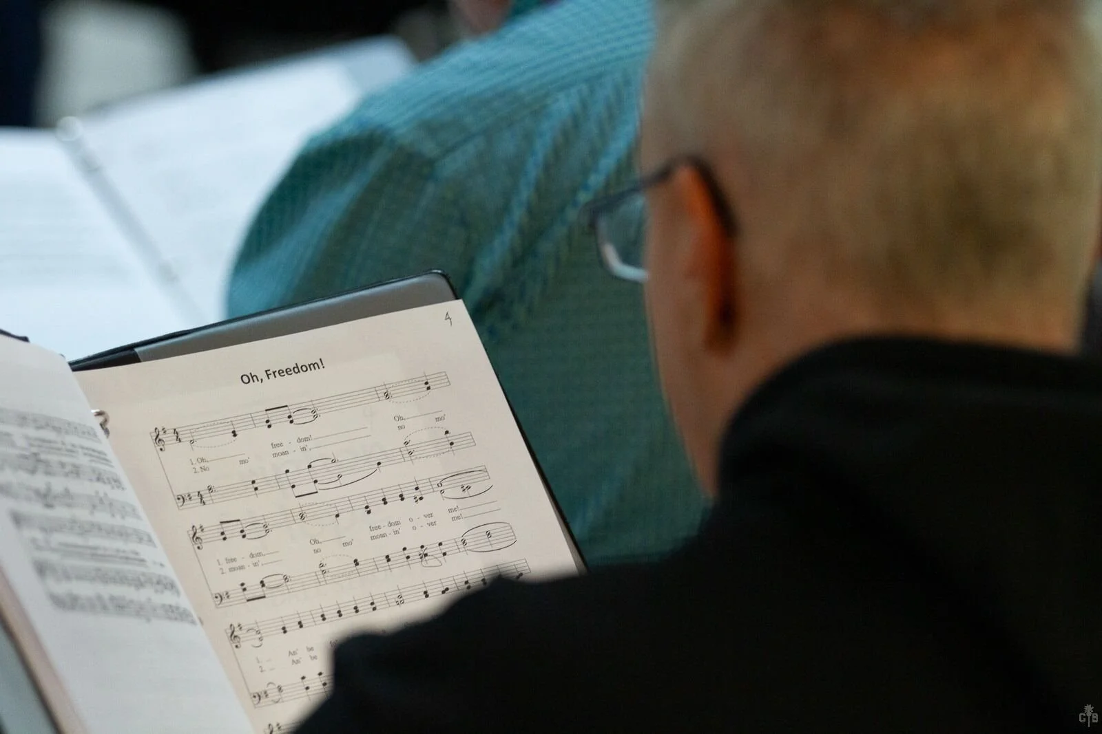 A man with glasses and short hair reading sheet music titled "Oh, Freedom!" in a conference or classroom setting.