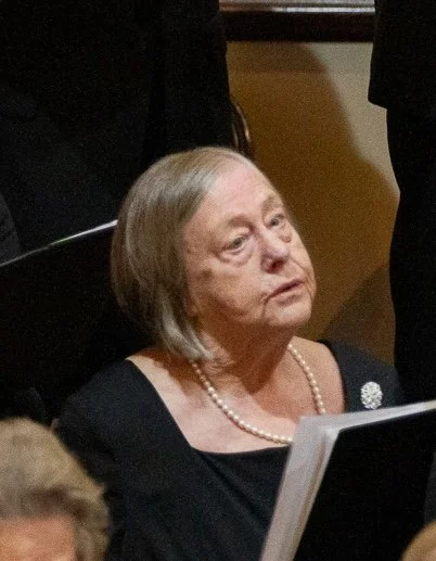 An elderly woman with gray hair wearing a black dress and pearl necklace, sitting in a formal setting.