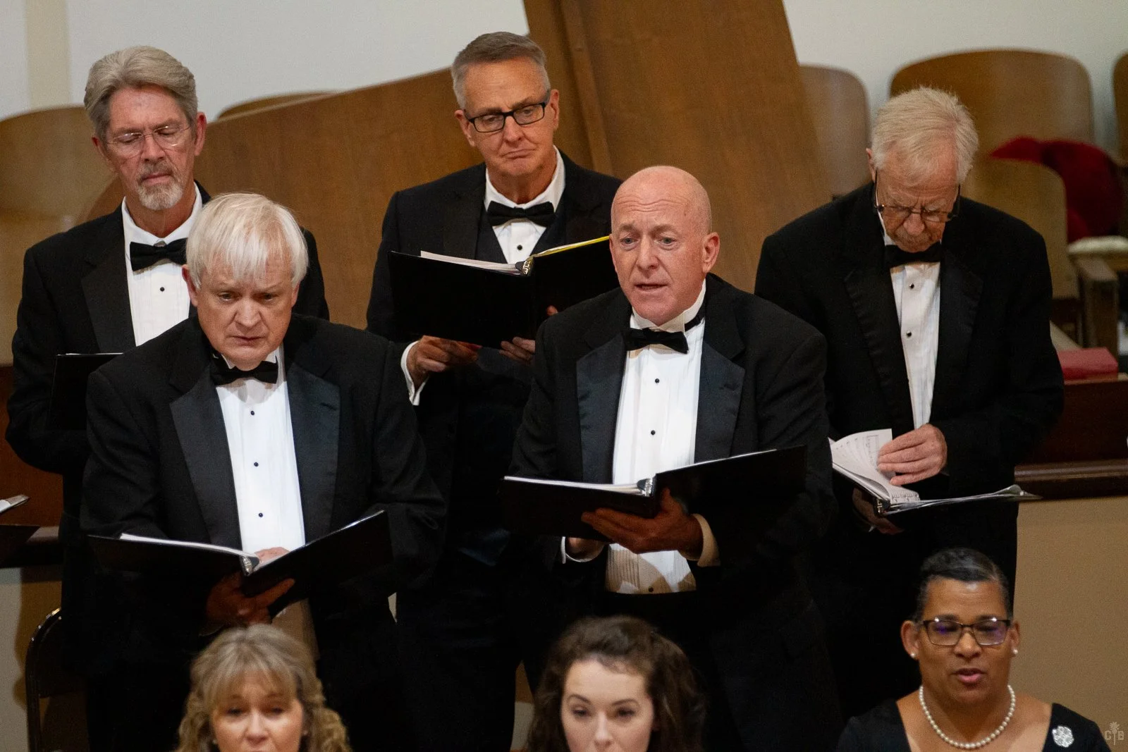 Five men in tuxedos singing from sheet music during a choir performance, seated in chairs in a concert hall.