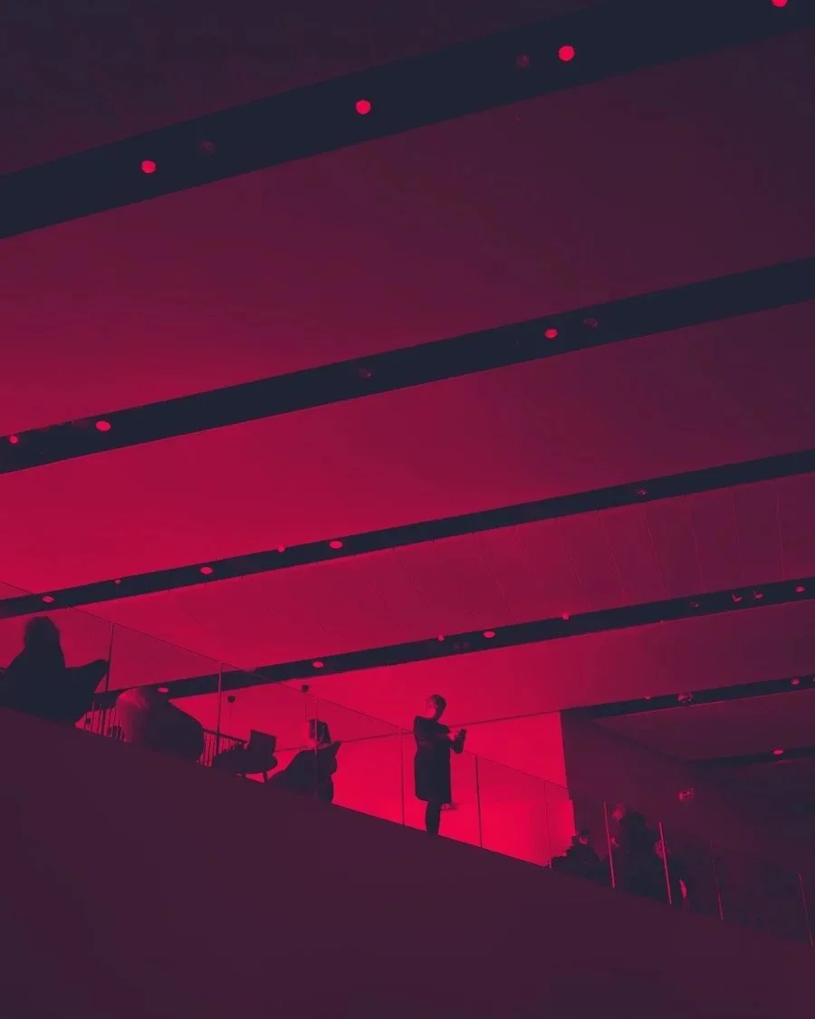 People standing on a balcony with red lighting, some using phones or tablets, in a modern interior space.