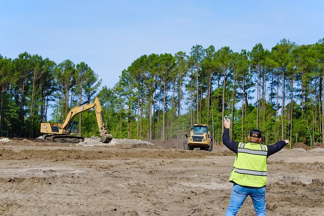 A construction site with a worker directing heavy machinery like an excavator and a compact loader, surrounded by trees and a clear blue sky.