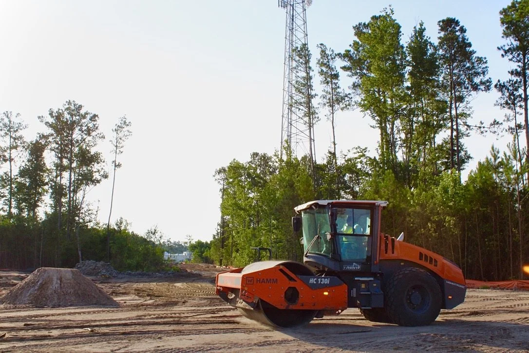 A steamroller compacting soil on a construction site during daytime, with trees and a communication tower in the background.