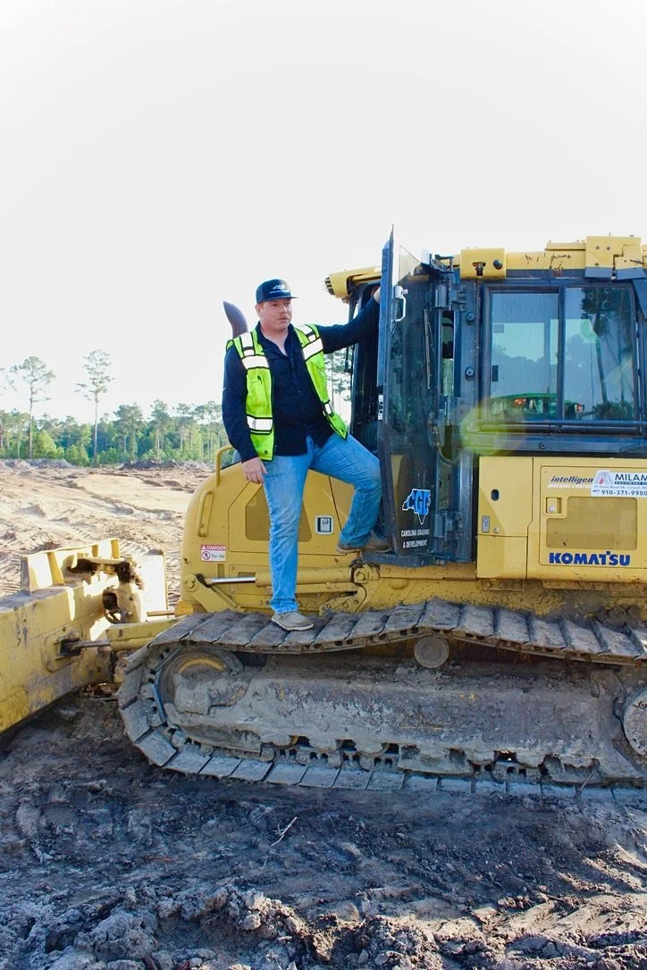 A man in a black cap, navy shirt, bright yellow safety vest, and light blue jeans stands on the track of a yellow Komatsu bulldozer at a construction site with dirt and trees in the background.