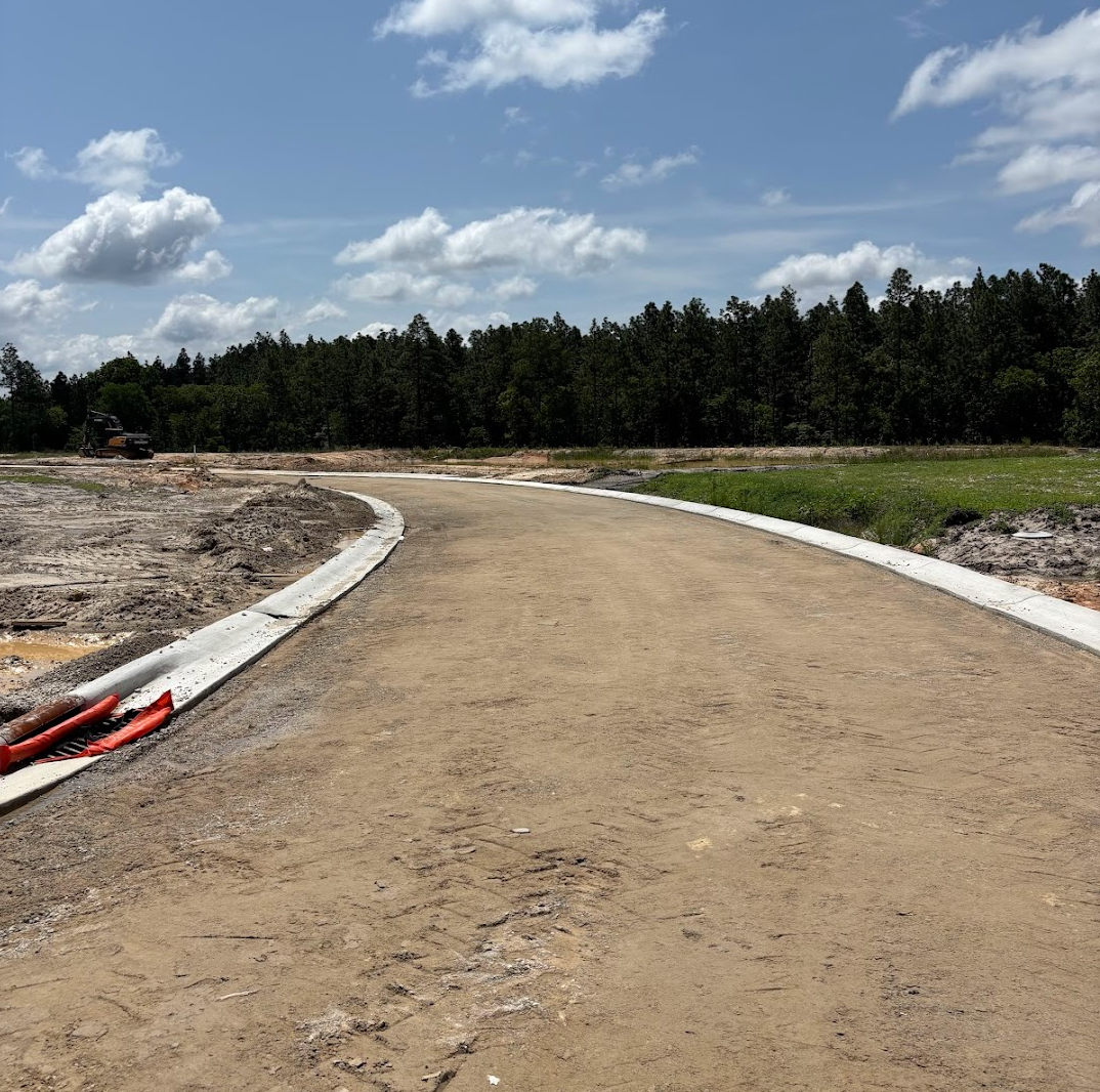 Unfinished paved road on a construction site with orange construction pipes on the side, surrounded by trees and blue sky with clouds.