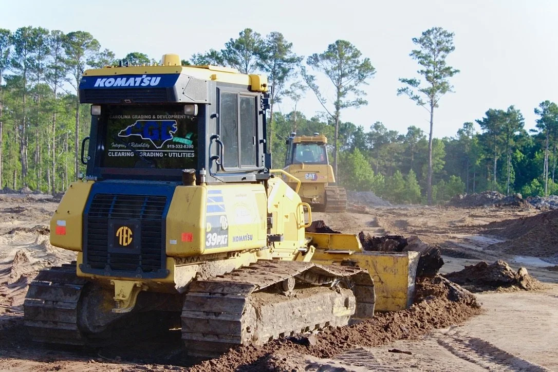 Construction site with heavy machinery, a yellow Komatsu bulldozer in the foreground and a yellow Caterpillar excavator in the background, surrounded by dirt and trees.