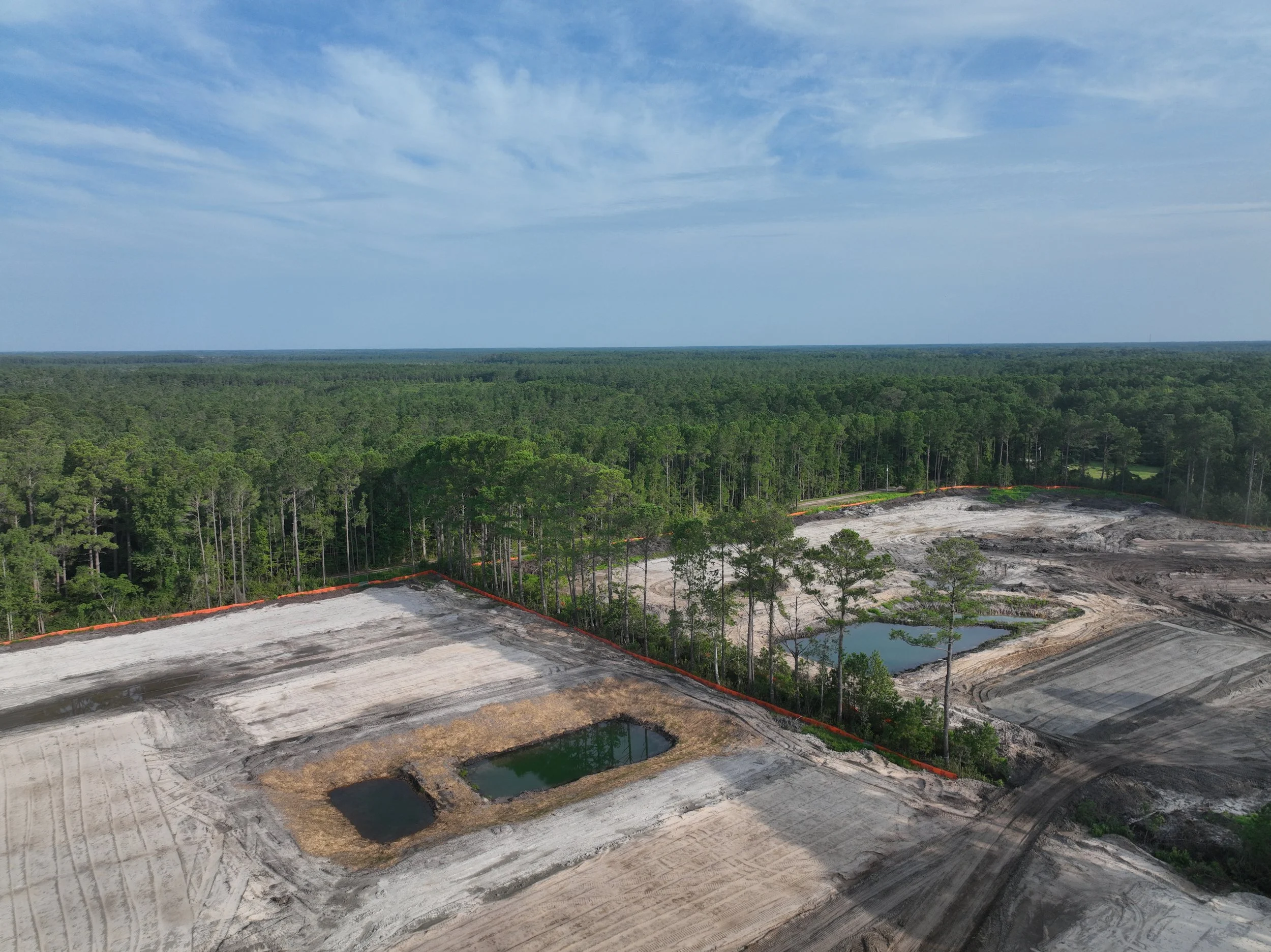 Construction site in a forested area with small ponds and cleared land under a partly cloudy sky.