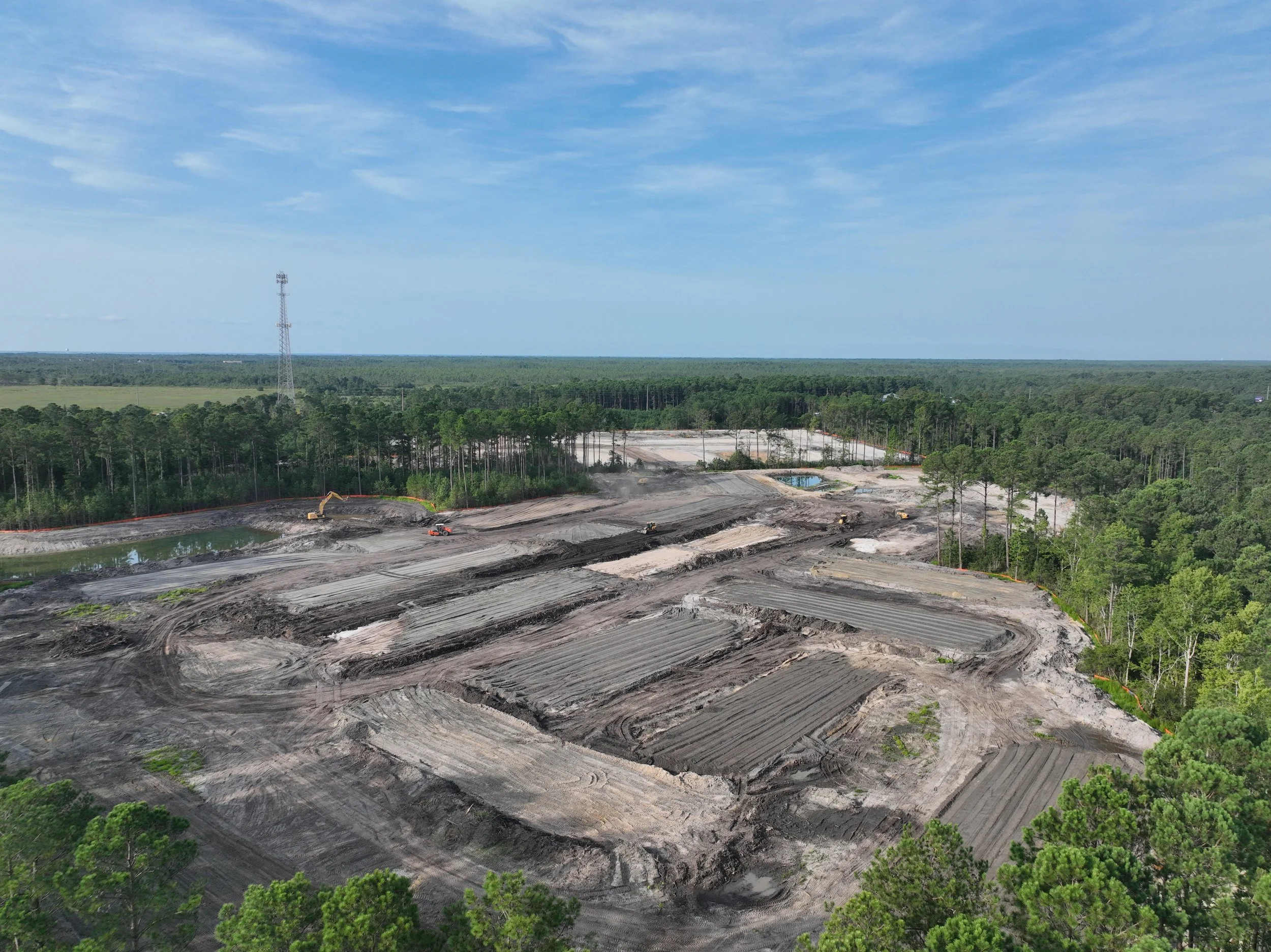 A wide view of a construction or land clearing site surrounded by a dense forest with a large open area showing earthwork, machinery, and a small water pond under a partly cloudy sky.