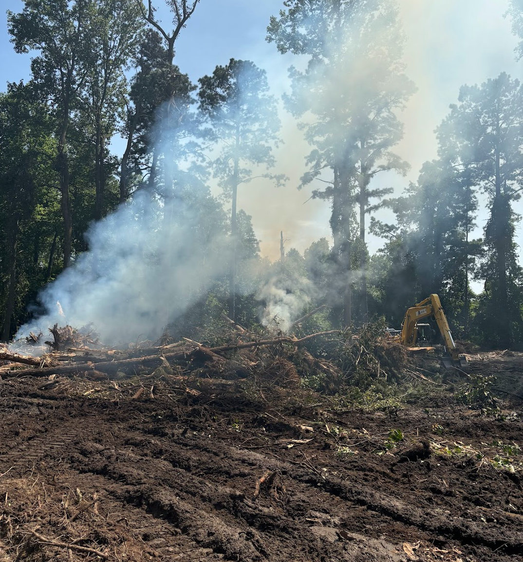 A hillside with fallen trees and smoke, a yellow excavator working on clearing the debris in a forested area.