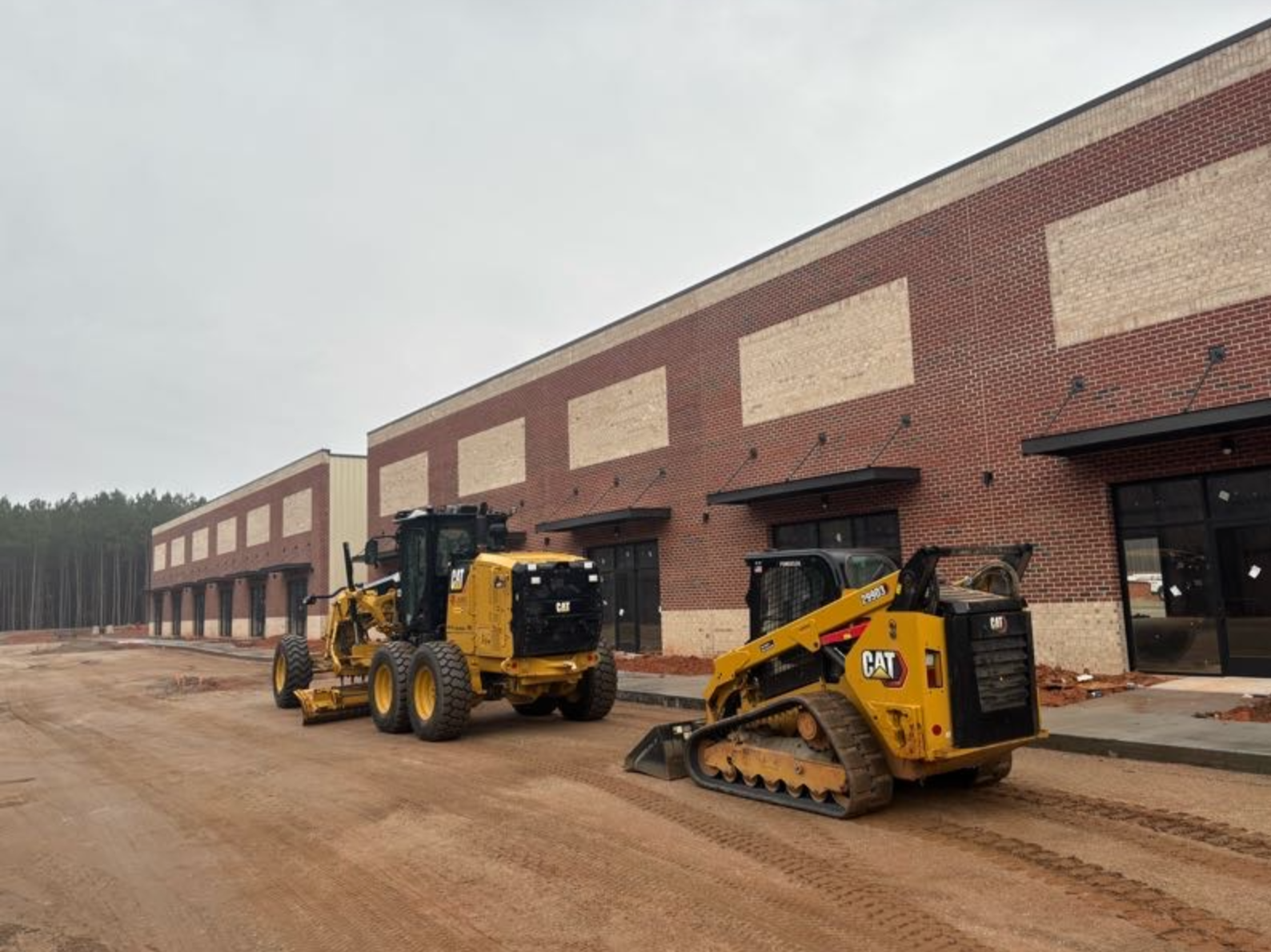 Construction site with a brick building under construction, two yellow Caterpillar vehicles on dirt ground, and unfinished storefronts with black awnings.