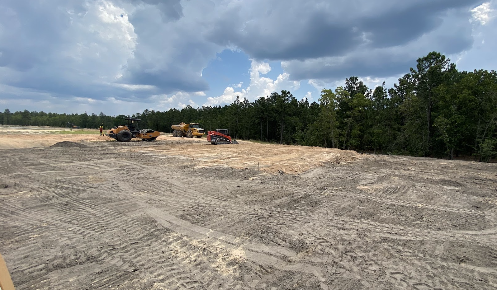 Construction site with machinery, dirt, and construction workers under a partly cloudy sky, surrounded by trees.