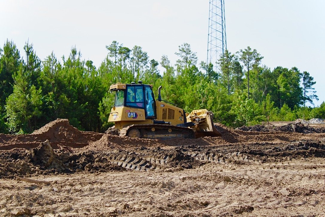 Construction site with a yellow Caterpillar bulldozer moving dirt, surrounded by trees and a tall radio tower in the background.