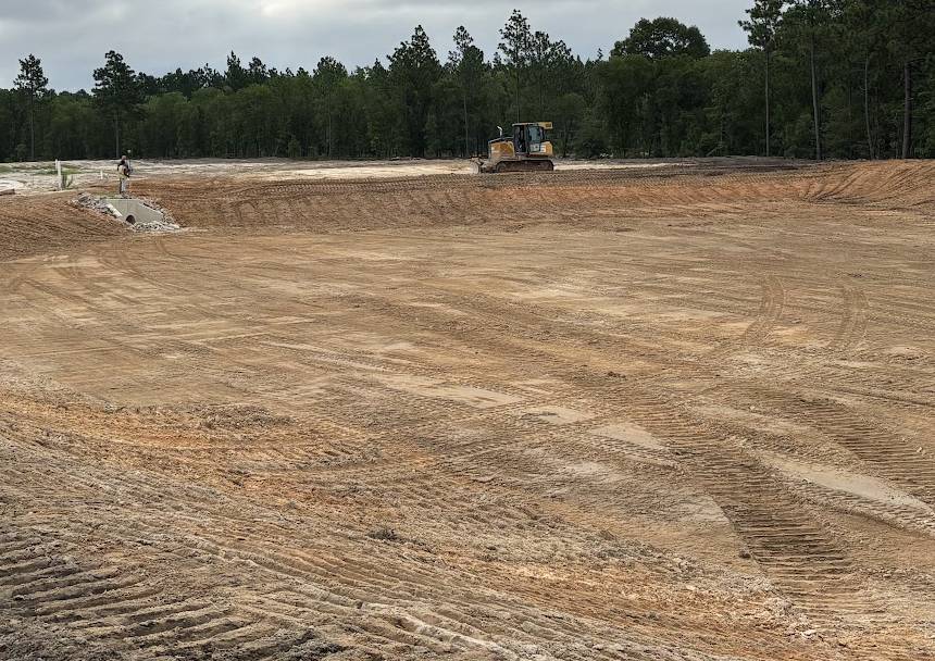 A construction site with a large flat area of dirt, tire tracks, and a bulldozer in the background, with trees and cloudy sky in the distance.