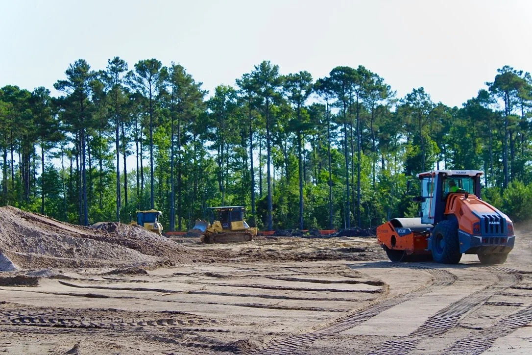 Construction site with bulldozer and two other pieces of heavy machinery working on dirt with trees in the background.