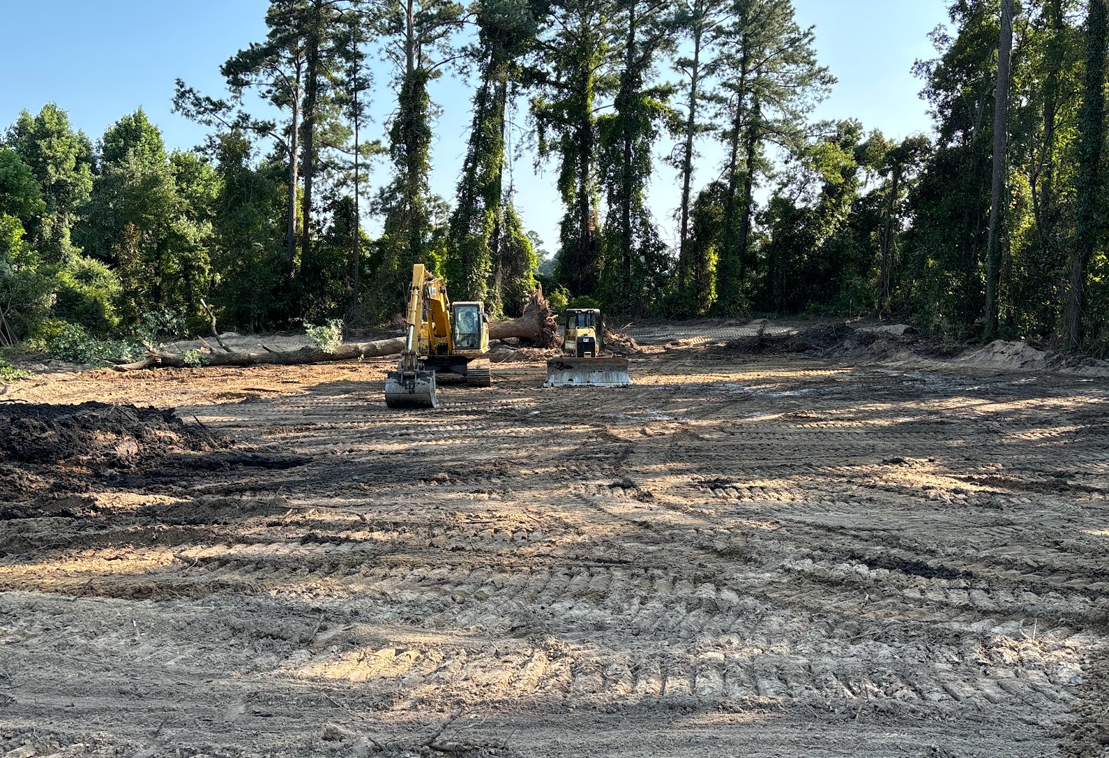 Construction site in a forest clearing with two excavators and a bulldozer on bare ground, surrounded by tall trees.