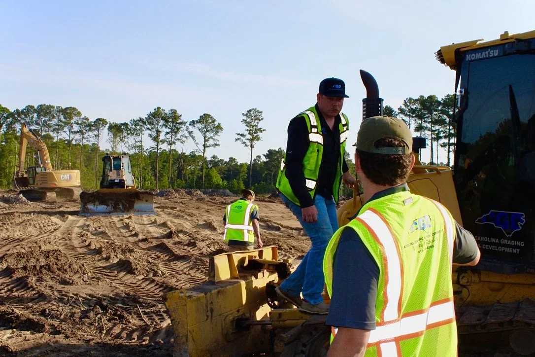 Construction workers operating heavy machinery on a dirt construction site, with trees in the background.