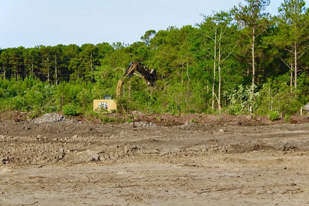 Construction site with excavator digging in a cleared area, surrounded by green trees and a clear sky.