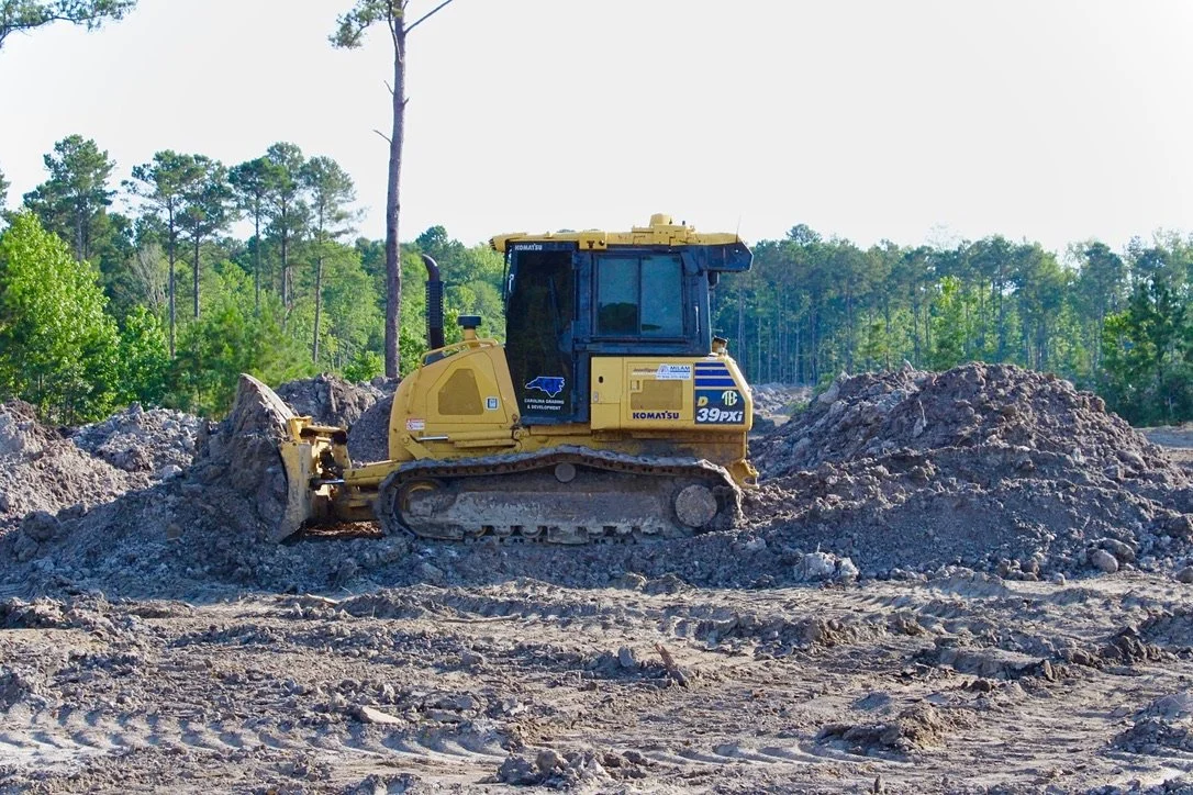 A yellow bulldozer working on a construction site with dirt and piles of soil, surrounded by a forest of green trees.