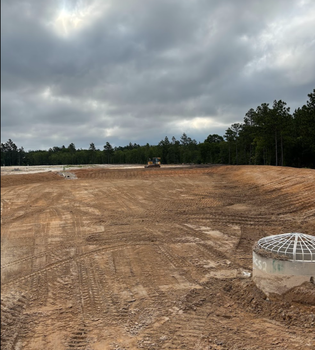 Construction site with dirt and tire tracks, a bulldozer in the distance, overcast sky, trees in the background.