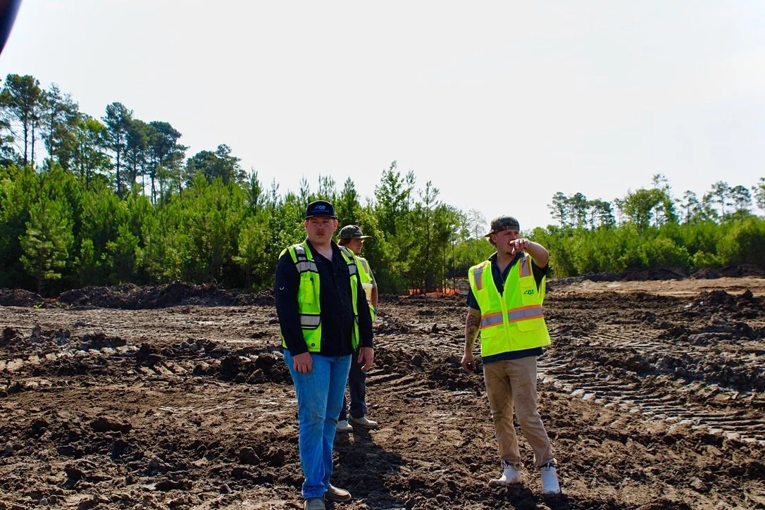Three construction workers in safety vests standing on a dirt construction site surrounded by trees.