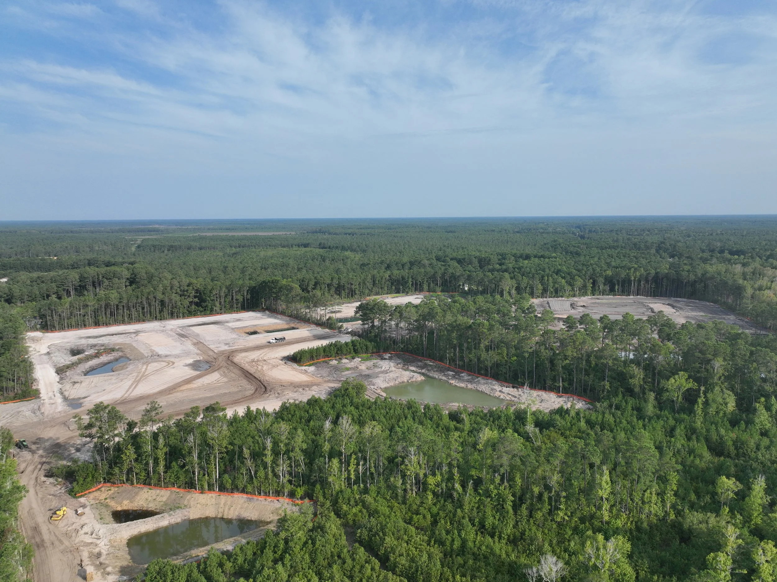 Aerial view of a forest with a cleared construction site that has some water bodies, machinery, and vehicles, with a blue sky with scattered clouds in the background.
