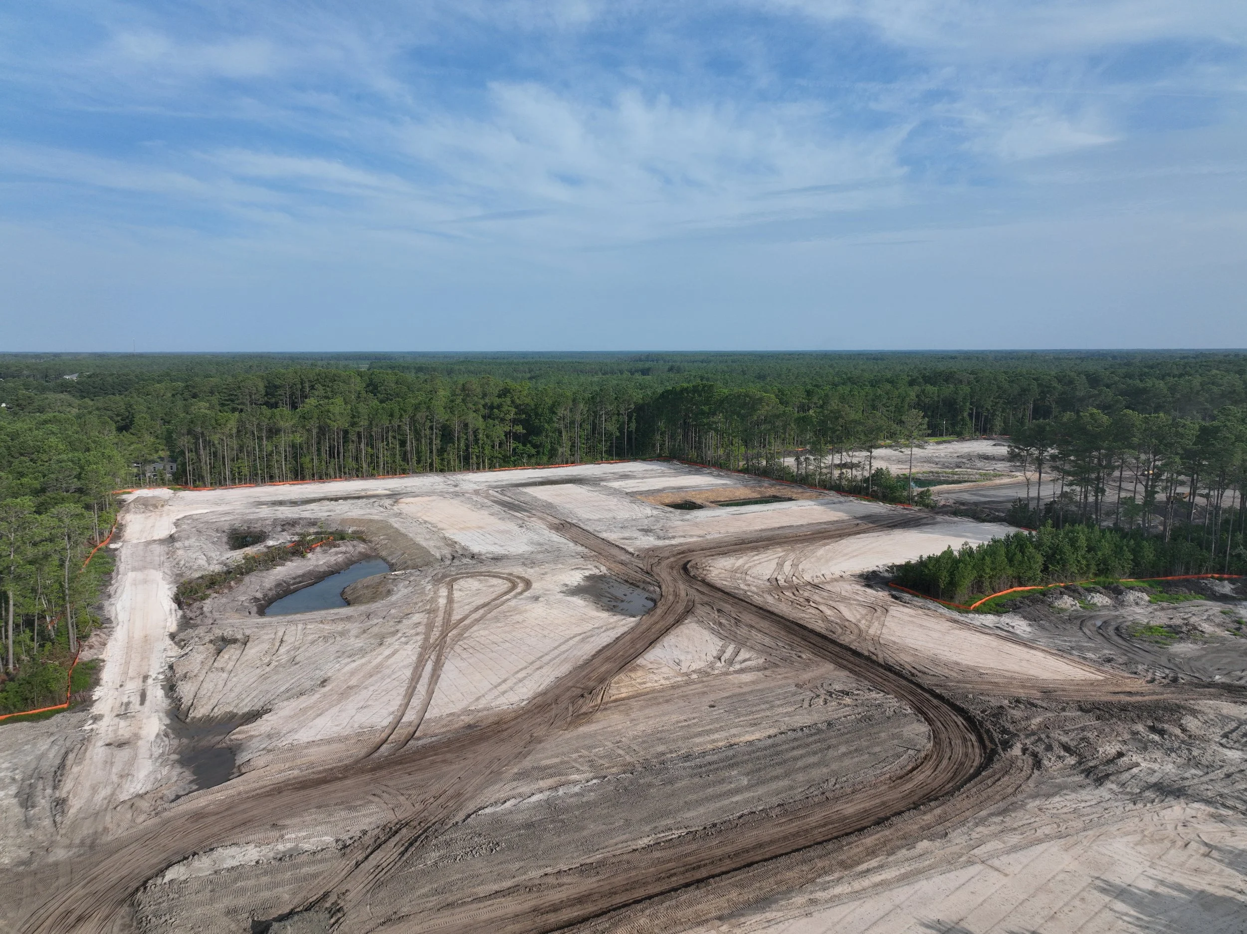 A landscape view of a construction site in a forested area with cleared land, water ponds, and patches of dirt, under a partly cloudy sky.