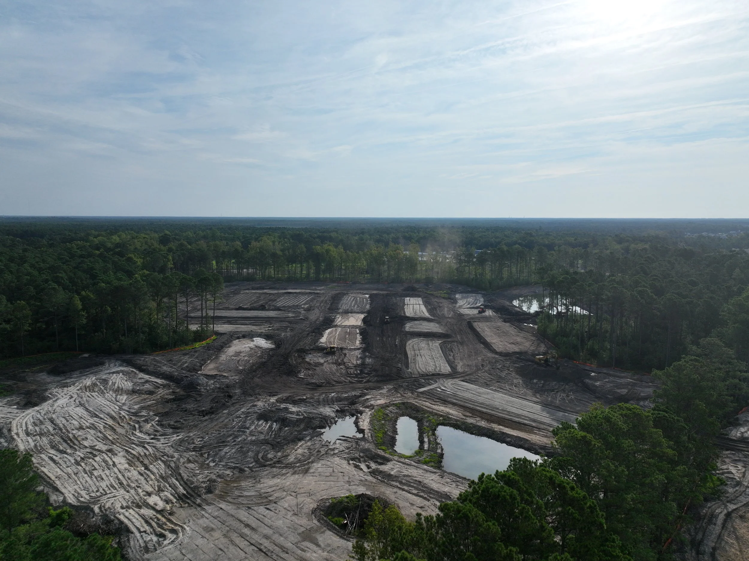 Aerial view of a landscape showing a cleared area with dirt and water, surrounded by dense green forests and a cloudy sky.