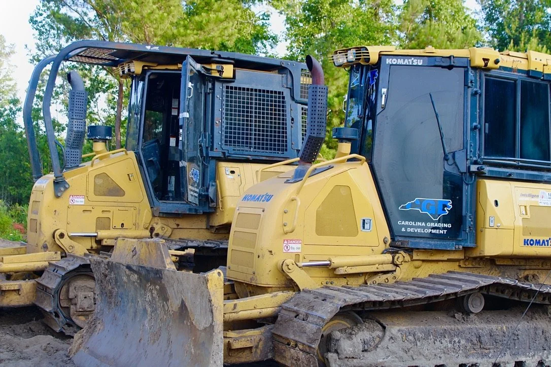 Two yellow Komatsu bulldozers on a construction site with green trees in the background.