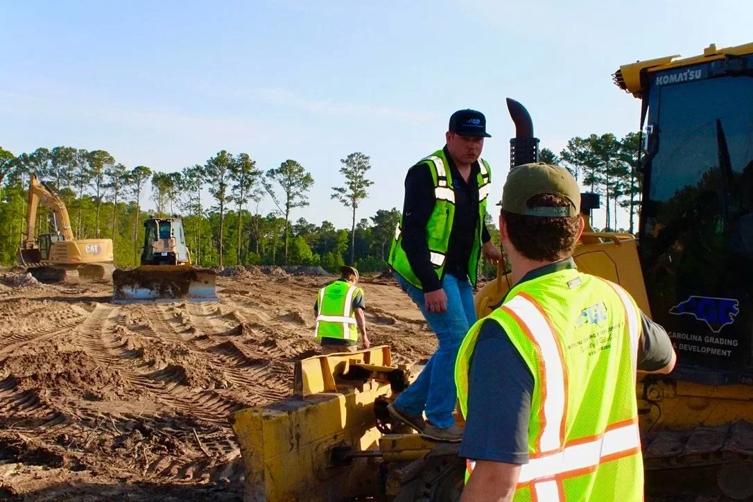 Construction workers wearing safety vests working on a construction site with heavy machinery and dirt terrain, with trees in the background.