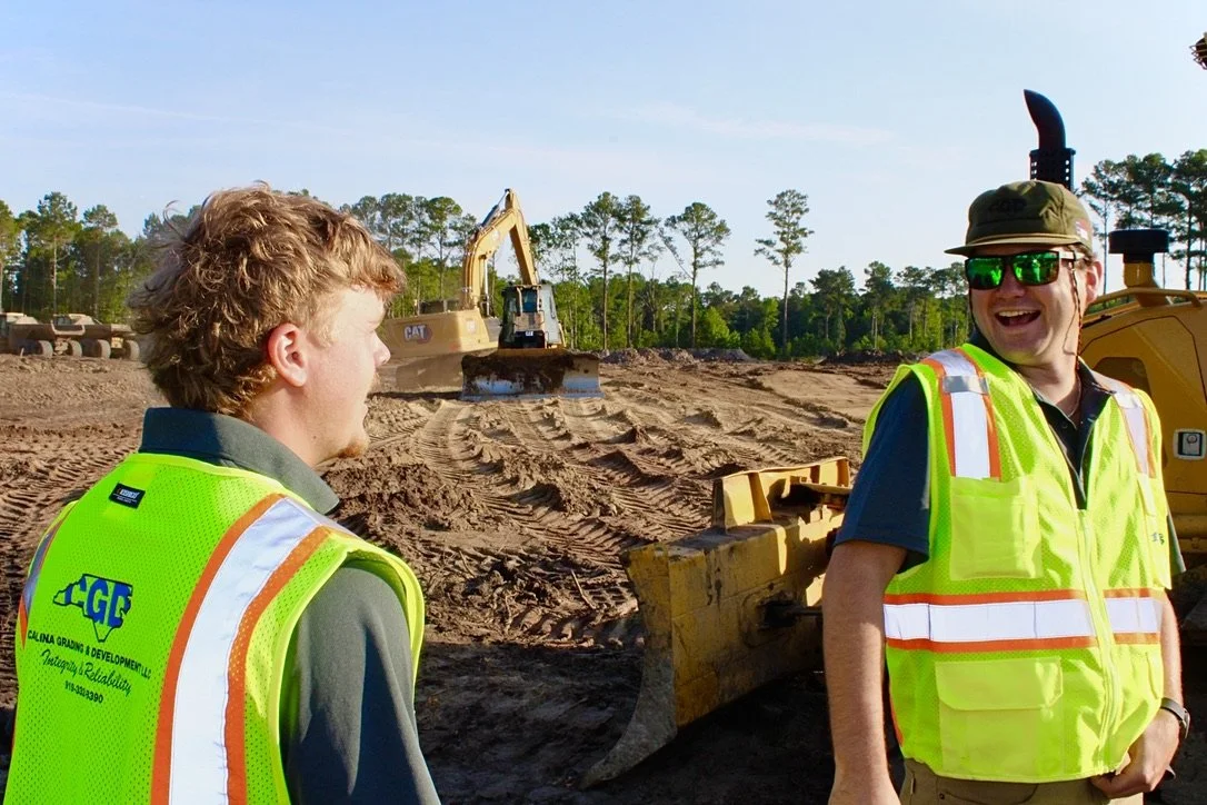 Two men wearing safety vests at a construction site, with heavy machinery and trees in the background.