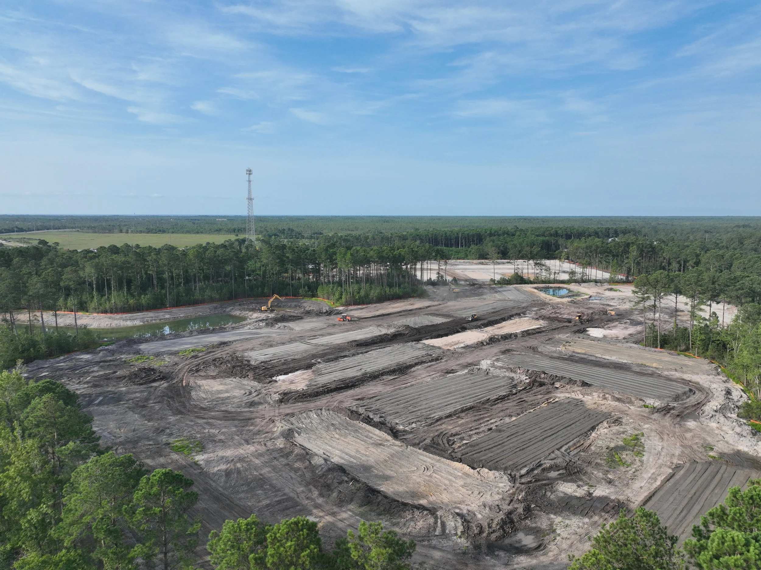 A construction site with dirt roads, construction equipment, surrounded by trees, and a distant landscape with a tall communication tower and open fields under a cloudy blue sky.