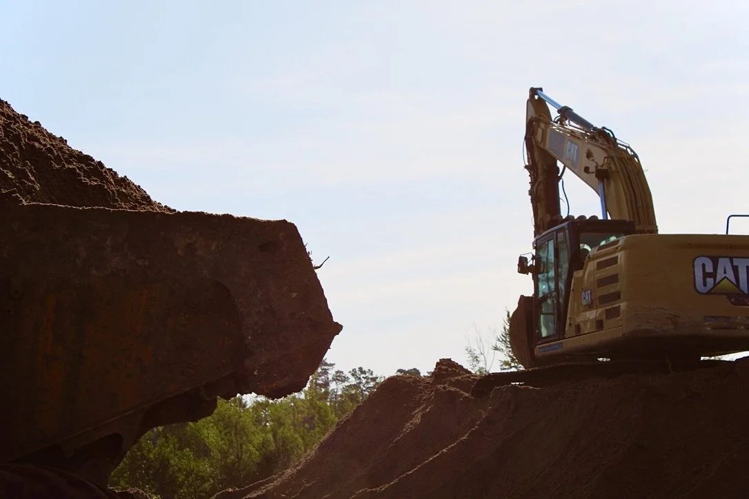 A yellow Caterpillar excavator on a mound of dirt at a construction site, with a large pile of dirt nearby and trees in the background under a clear sky.