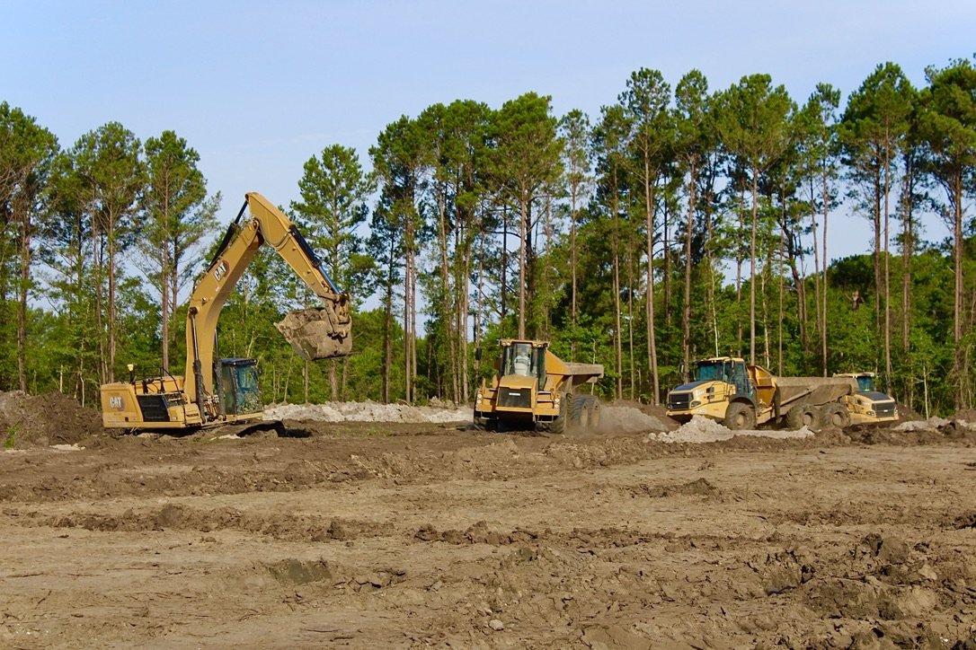 Construction site with three yellow heavy machinery vehicles working on leveling and excavating the dirt, with green trees in the background.